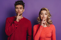 A man and a woman stand side by side against a purple background. Both have their index fingers placed over their lips, signaling for silence. The man wears a red sweater, and the woman wears a coral long-sleeve top. Both have a serious expression.