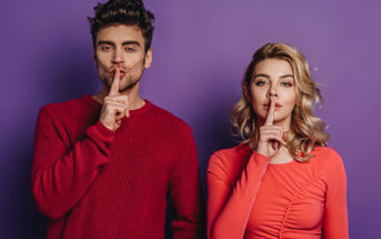 A man and a woman stand side by side against a purple background. Both have their index fingers placed over their lips, signaling for silence. The man wears a red sweater, and the woman wears a coral long-sleeve top. Both have a serious expression.