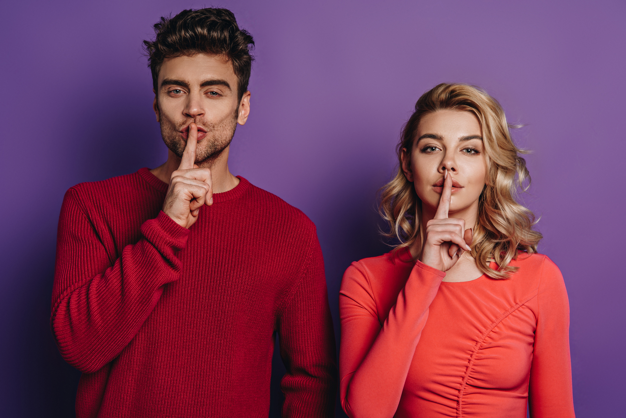 A man and a woman stand side by side against a purple background. Both have their index fingers placed over their lips, signaling for silence. The man wears a red sweater, and the woman wears a coral long-sleeve top. Both have a serious expression.