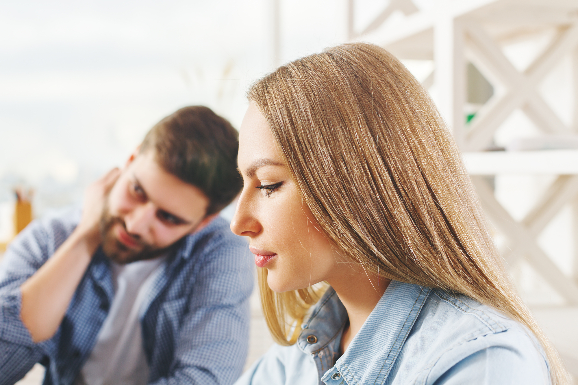 A woman with long hair is sitting and looking down thoughtfully, while a man sits behind her, glancing in her direction with a hand on his neck. Both appear to be indoors with a bright, softly lit background.