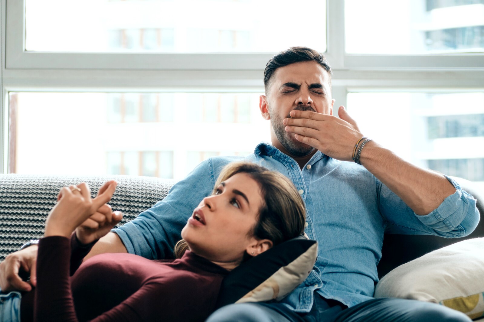 A man and woman are relaxing on a couch. The woman, in a maroon top, is lying with her head on the man's lap, pointing upwards seemingly explaining something. The man, in a blue shirt, is yawning with his hand covering his mouth. Light fills the room from large windows.