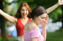 Two women are in an outdoor setting with trees in the background. One woman in a red top stands in the background with arms raised, while the woman in the foreground, wearing a pink off-shoulder top, appears upset or uncomfortable, with her hand near her face.