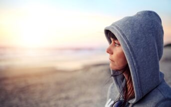 A person wearing a gray hoodie stands on a beach, gazing thoughtfully towards the horizon during sunset. The sandy beach and calm ocean can be seen in the background, bathed in soft, warm light.