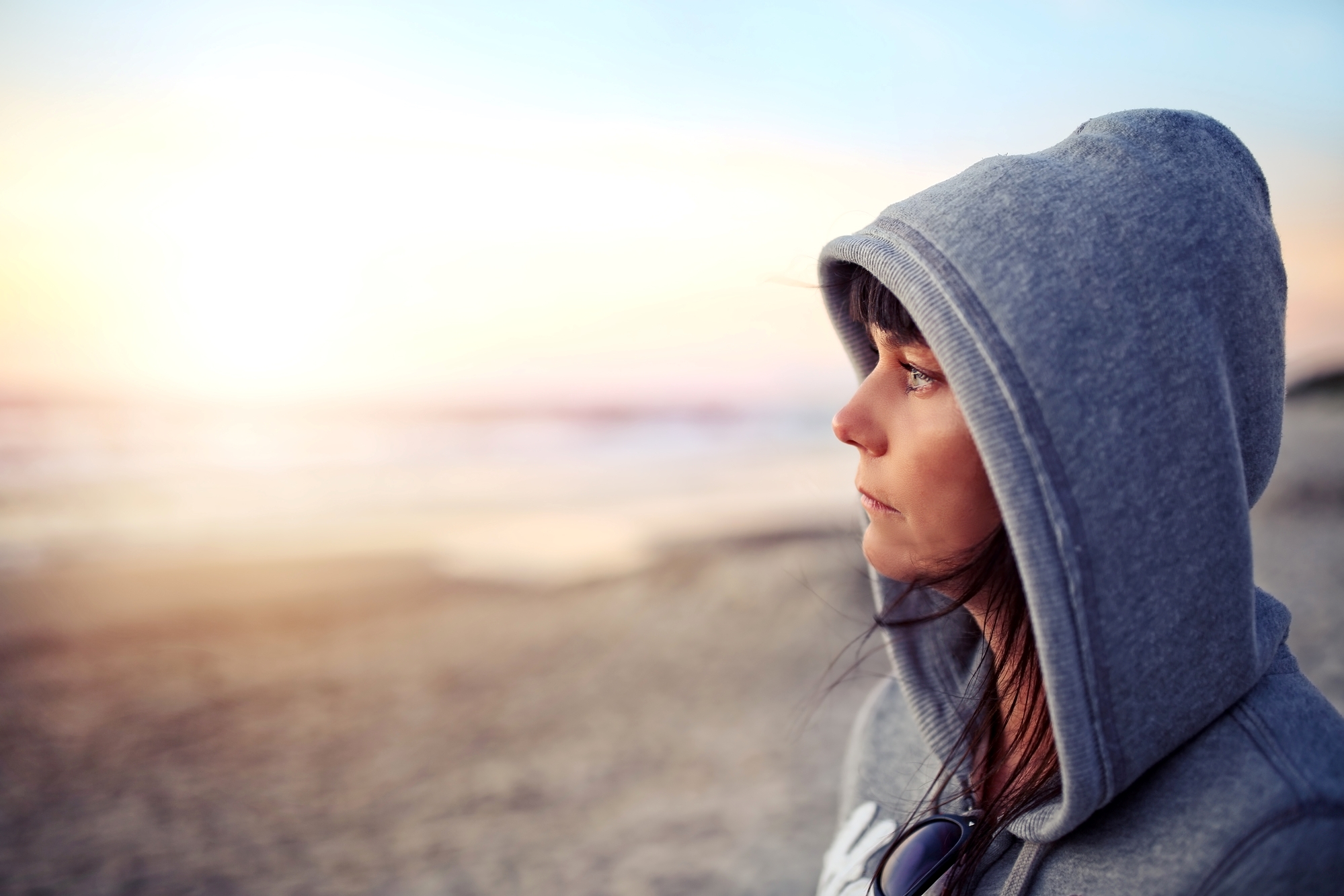 A person wearing a gray hoodie stands on a beach, gazing thoughtfully towards the horizon during sunset. The sandy beach and calm ocean can be seen in the background, bathed in soft, warm light.