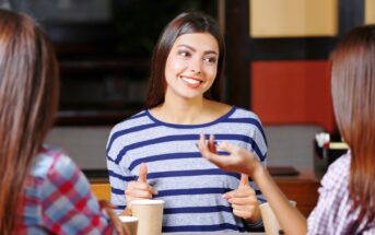 Three women enjoying a conversation at a coffee shop, with disposable coffee cups on the table. The woman in the center is wearing a striped shirt and smiling, actively engaging in the discussion. The other two are seen from behind, wearing colorful plaid shirts.