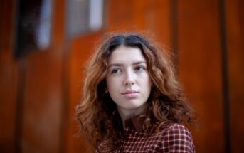 A person with wavy, shoulder-length brown hair wearing a maroon and white checkered shirt looks slightly to the side with a neutral expression. The background features a blurred, reddish-orange wall.