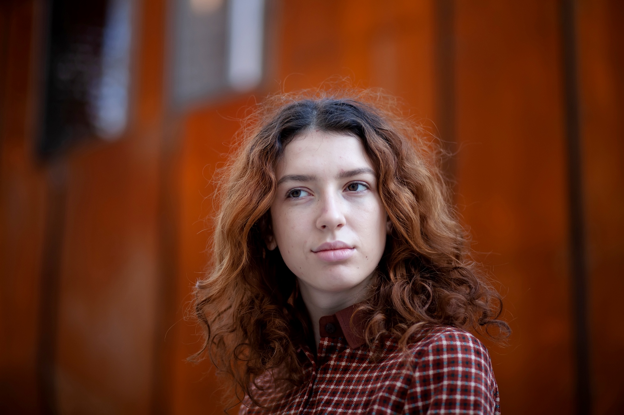 A person with wavy, shoulder-length brown hair wearing a maroon and white checkered shirt looks slightly to the side with a neutral expression. The background features a blurred, reddish-orange wall.