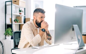 A bearded man sits at a desk, intently looking at a computer monitor. His hands are clasped near his mouth in concentration. He is in a bright office space with shelves, plants, and other items in the background.