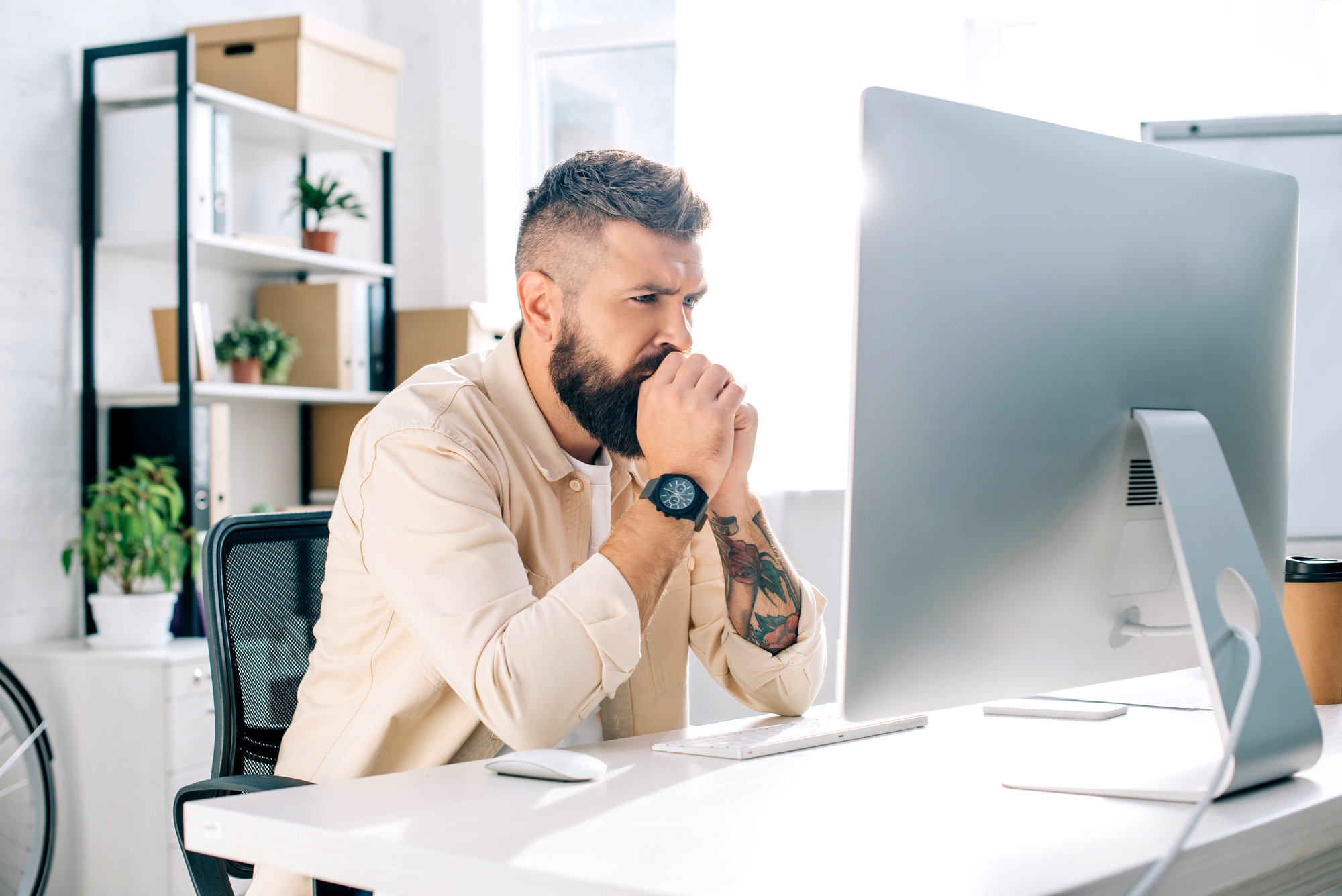 A bearded man sits at a desk, intently looking at a computer monitor. His hands are clasped near his mouth in concentration. He is in a bright office space with shelves, plants, and other items in the background.