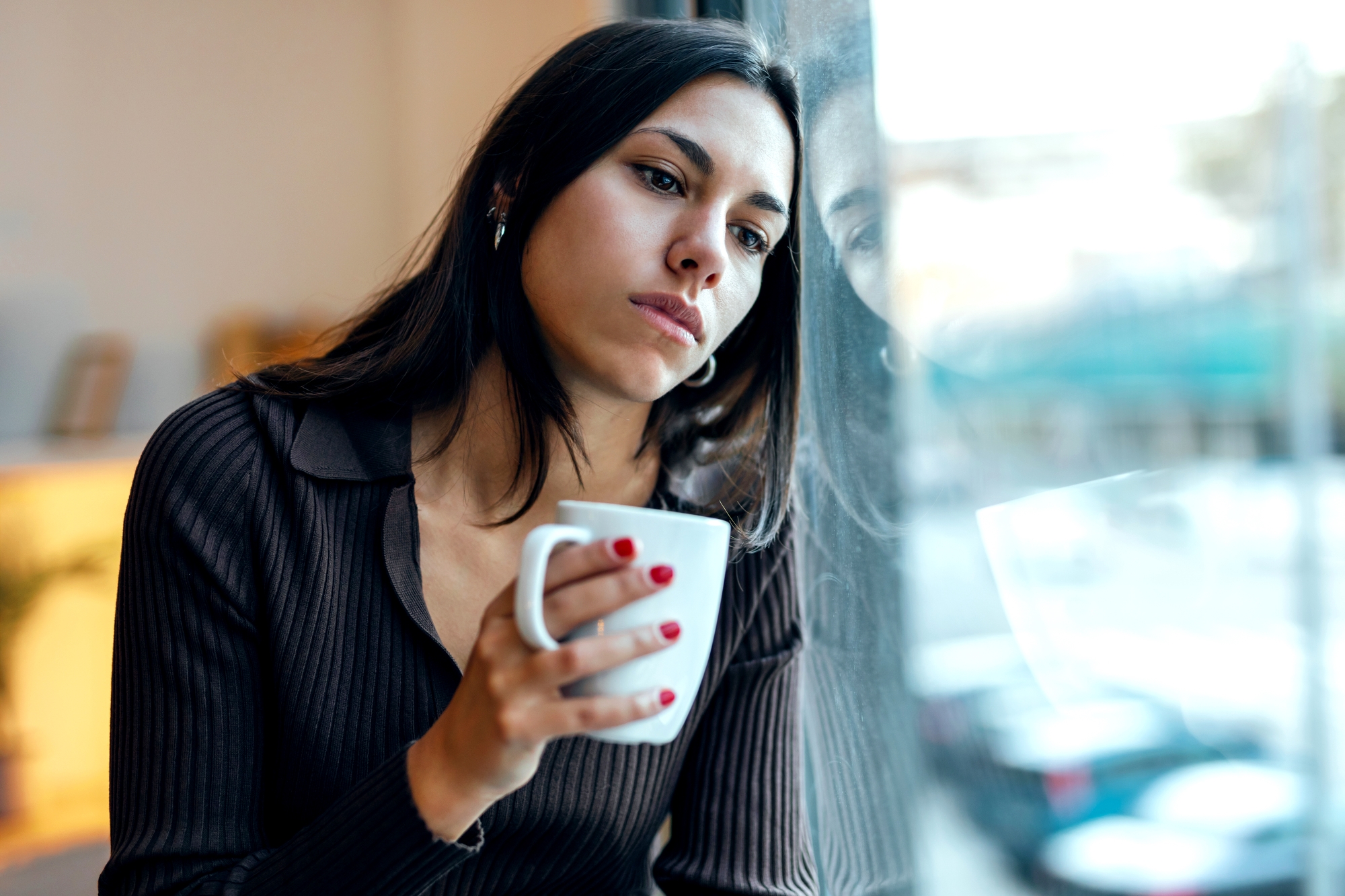 A woman with long dark hair sits by a window, holding a white mug. She looks contemplative and appears to be lost in thought, gazing out of the window. She is wearing a dark, long-sleeved top, and her fingernails are painted red.