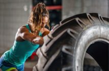 A woman in athletic wear is flipping a large tire in a gym. She has her hair tied back and is focused on the task. The background shows gym equipment and a tiled wall. The lighting highlights her muscles and determination.