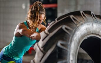 A woman in athletic wear is flipping a large tire in a gym. She has her hair tied back and is focused on the task. The background shows gym equipment and a tiled wall. The lighting highlights her muscles and determination.