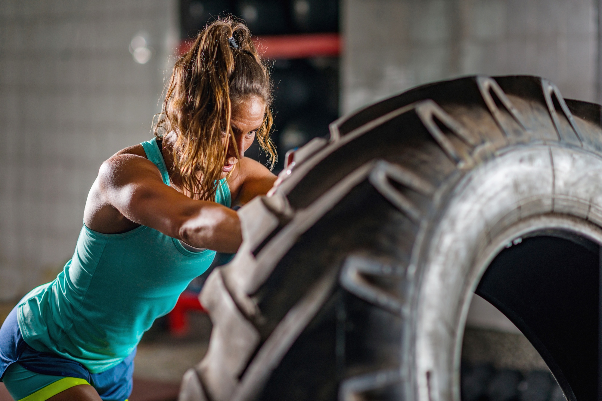 A woman in athletic wear is flipping a large tire in a gym. She has her hair tied back and is focused on the task. The background shows gym equipment and a tiled wall. The lighting highlights her muscles and determination.