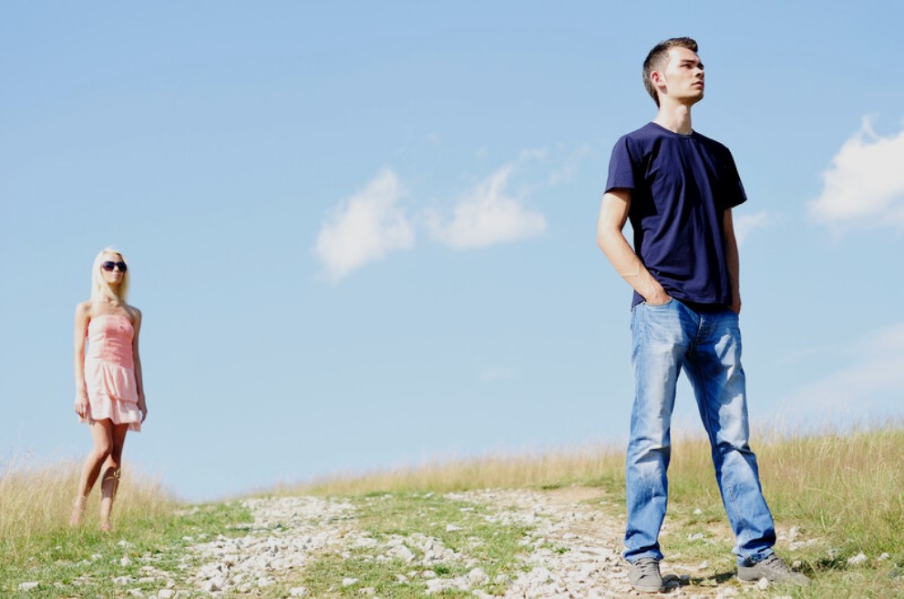 A man in a navy t-shirt and jeans stands with his hands in his pockets, looking to the right. A woman in a light pink dress and sunglasses stands in the background. They are in an open grassy field under a clear blue sky.