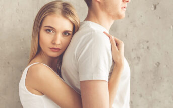 A young woman with long blonde hair, wearing a white tank top, stands behind a man in a white t-shirt, resting her head on his back and wrapping her arms around him. Both are standing against a textured gray wall, looking serious.