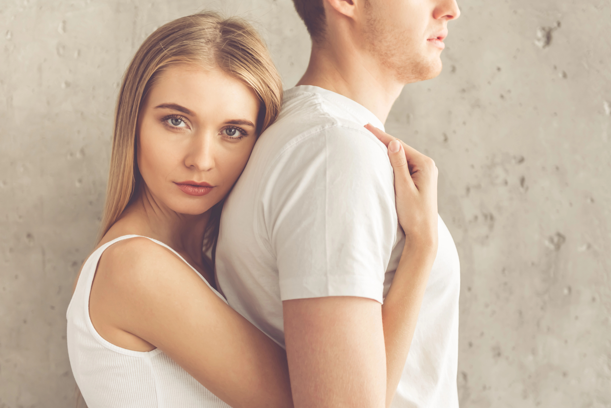 A young woman with long blonde hair, wearing a white tank top, stands behind a man in a white t-shirt, resting her head on his back and wrapping her arms around him. Both are standing against a textured gray wall, looking serious.