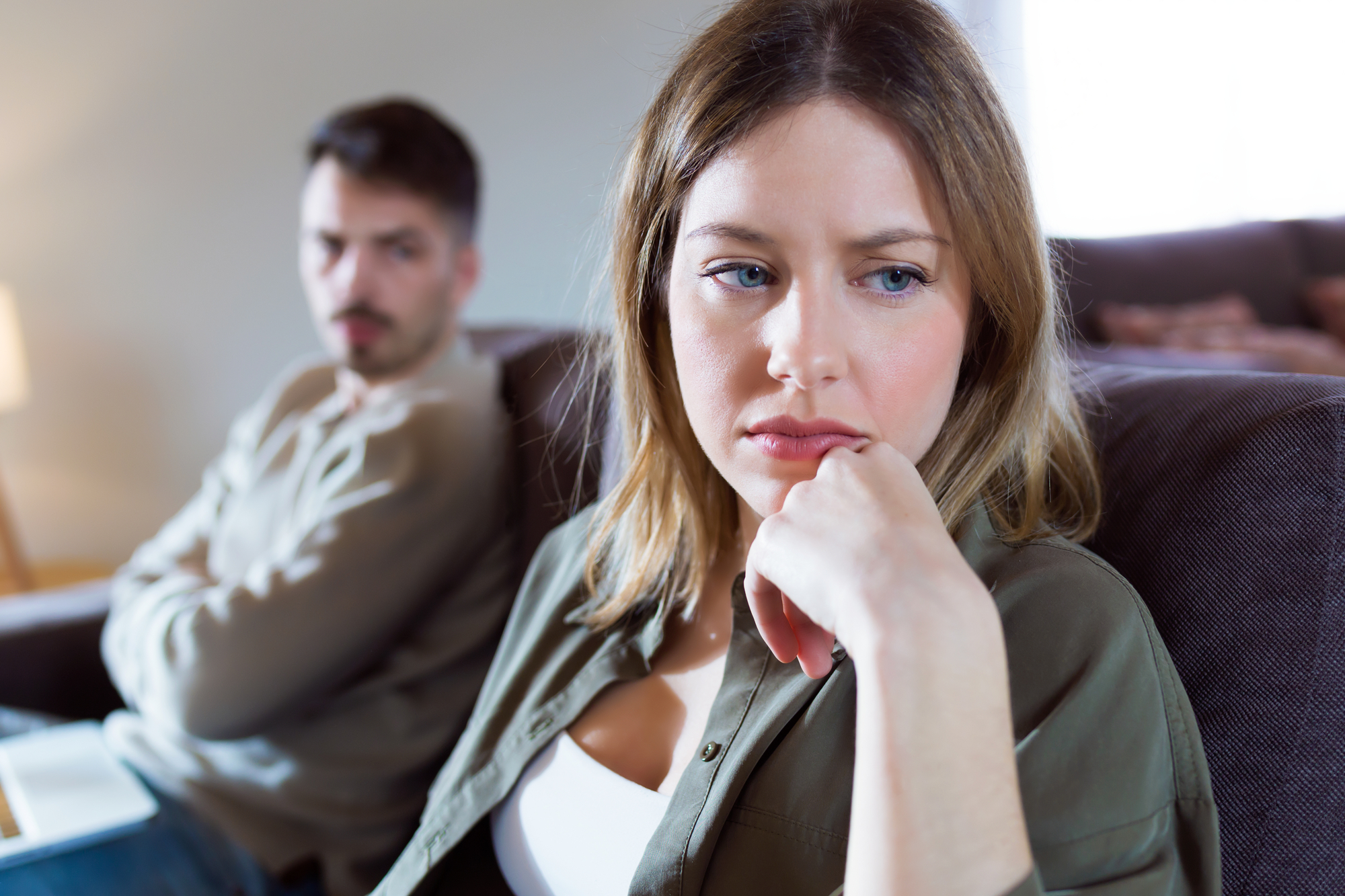 A woman with blonde hair sits on a couch, looking thoughtful and slightly distressed, with her hand resting on her chin. In the background, a man with a beard sits with his arms crossed, gazing towards her with a serious expression. The setting appears to be a living room.