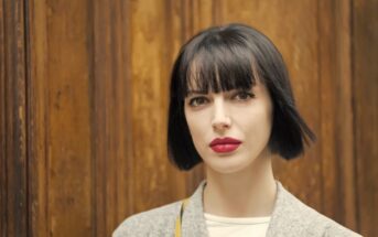 A person with short black hair and bangs stands in front of a wooden background. They are wearing a light-colored coat and white shirt, with noticeable red lipstick. The overall expression appears neutral.