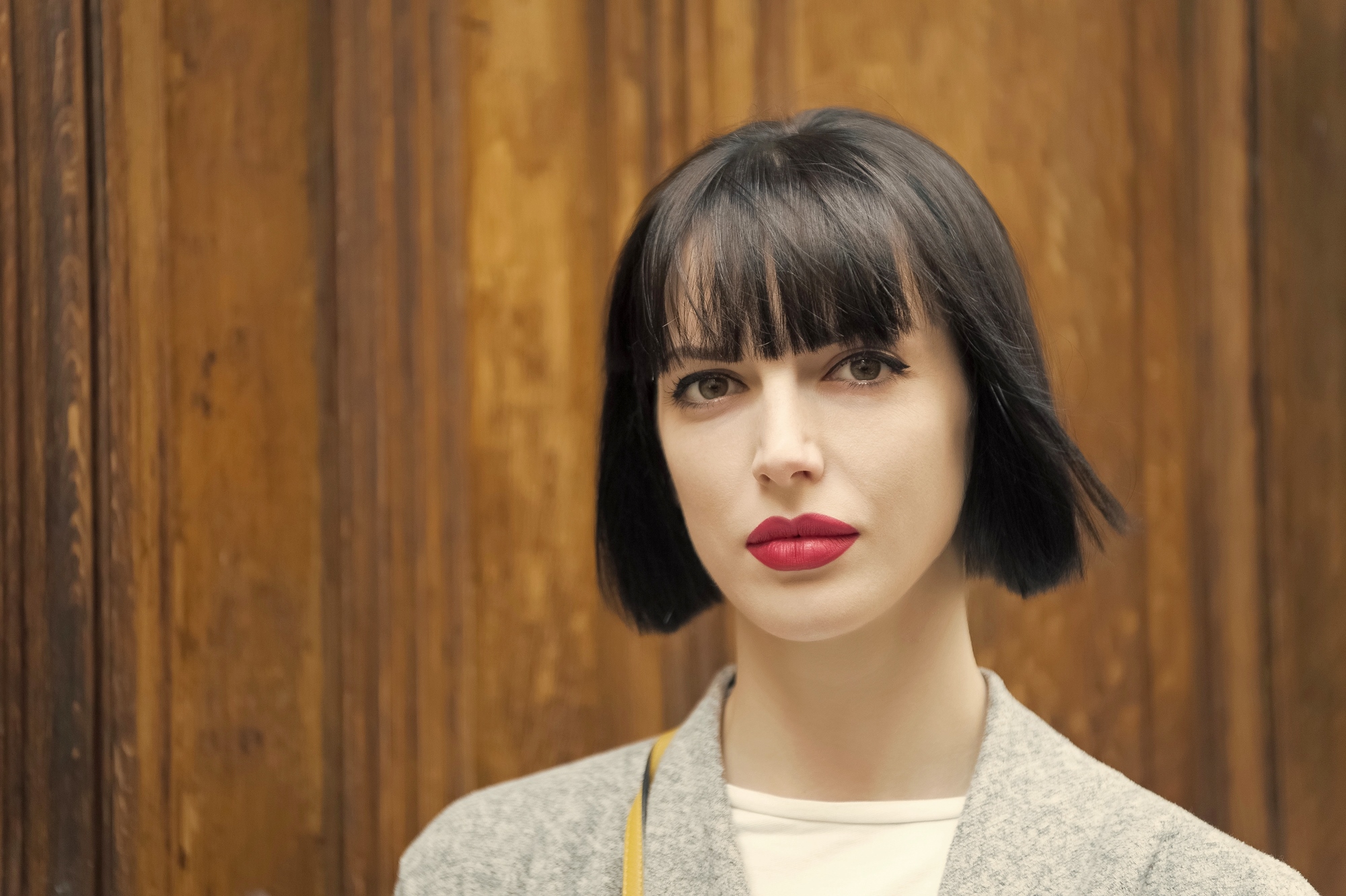 A person with short black hair and bangs stands in front of a wooden background. They are wearing a light-colored coat and white shirt, with noticeable red lipstick. The overall expression appears neutral.