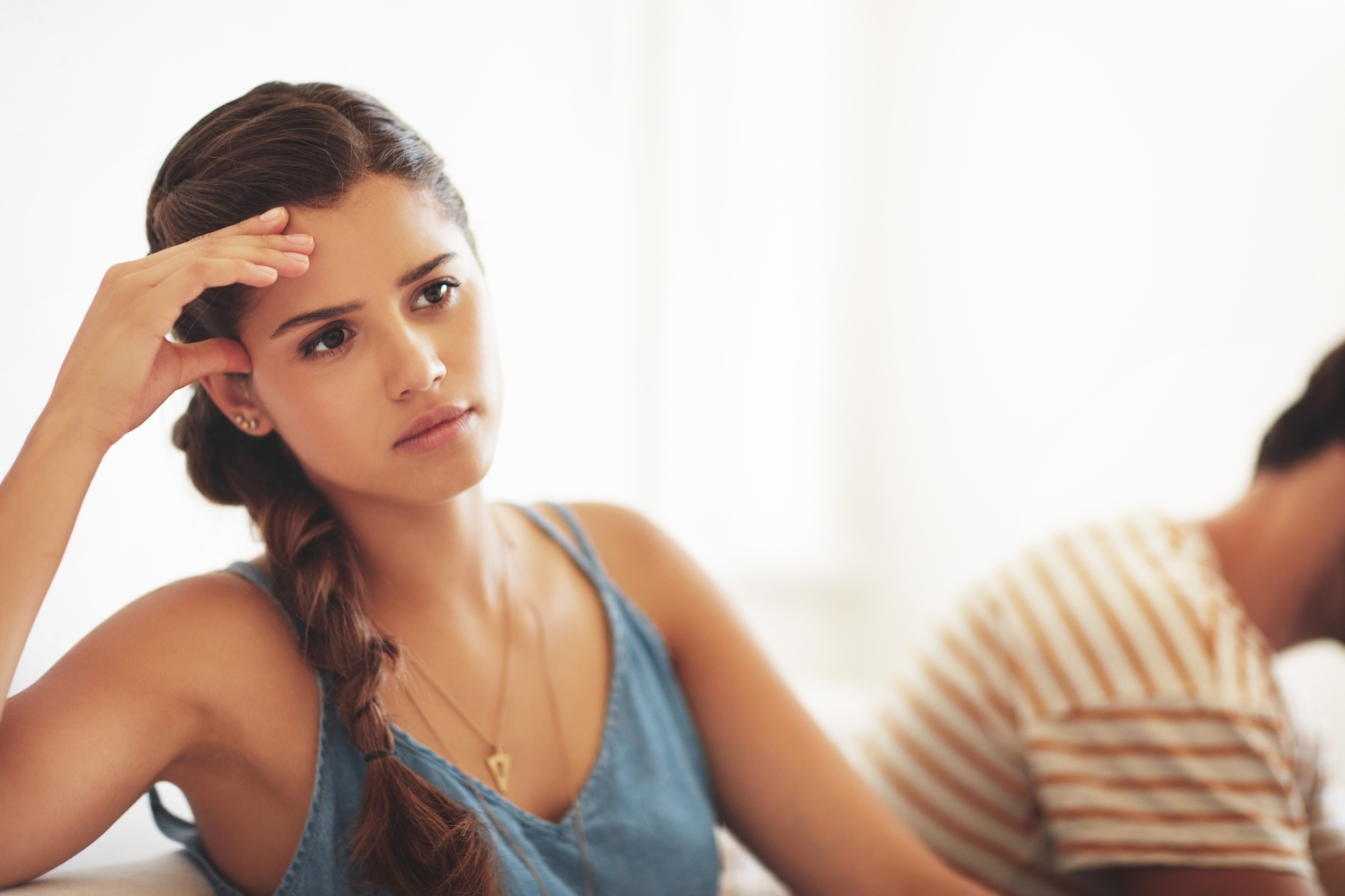 A woman with a long braid, wearing a blue sleeveless top, sits in the foreground with a thoughtful and concerned expression, resting her head on her hand. In the background, a person in a striped shirt is blurred and facing away. The background is light and minimalistic.