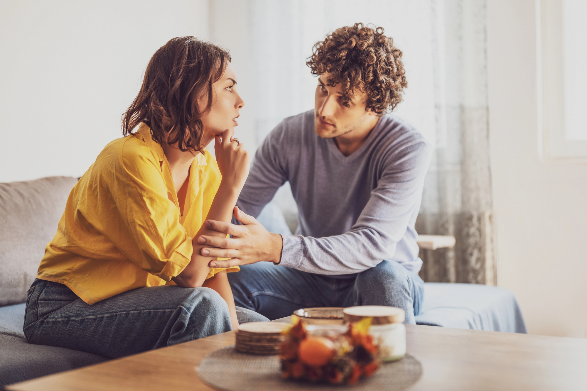 A man and woman sit closely on a couch in a conversation. The man, wearing a gray long-sleeve shirt and jeans, gently touches the woman's arm. The woman, dressed in a yellow shirt and jeans, listens intently, her hand under her chin. The room is well-lit and cozy.