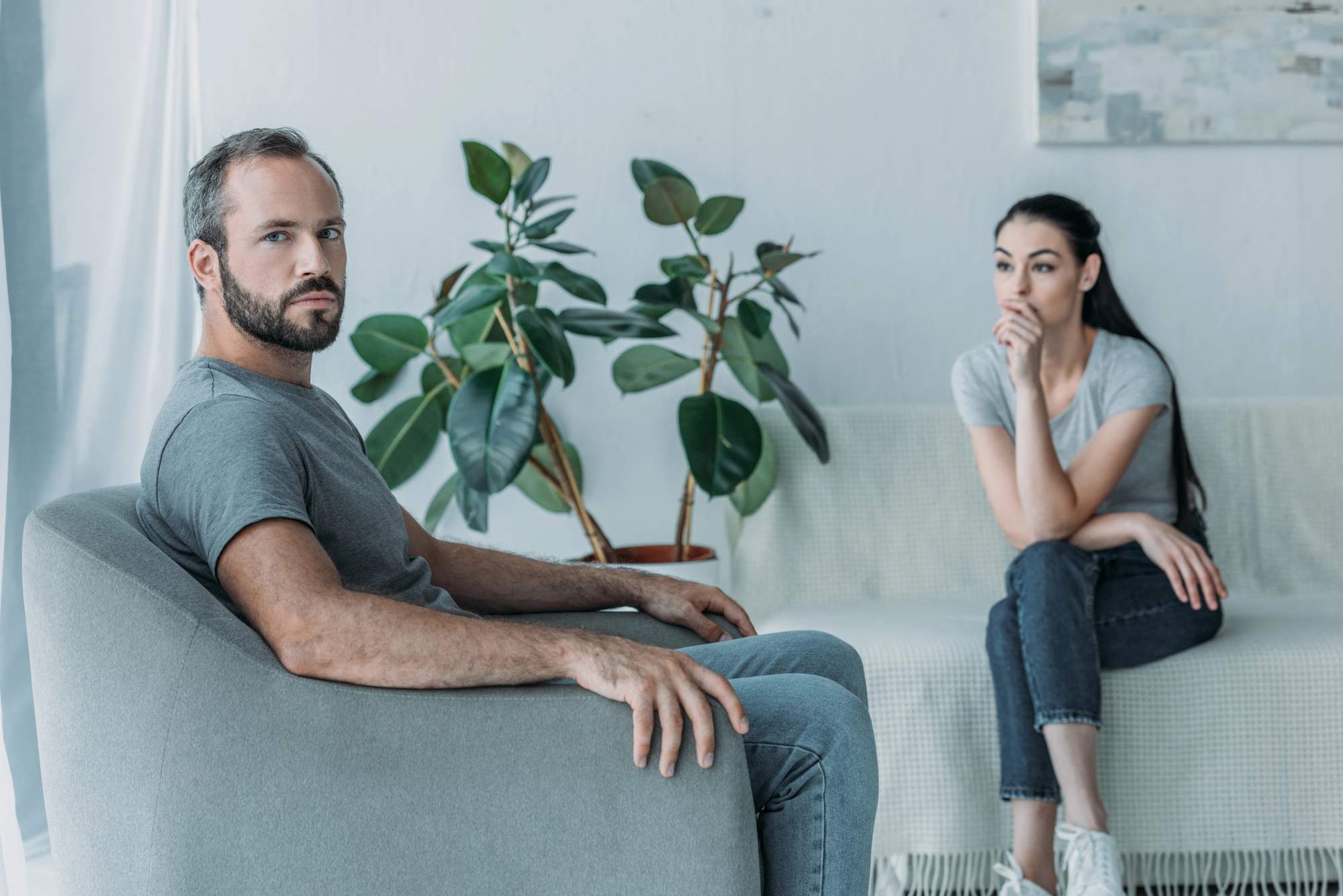 A man and a woman are sitting in a living room. The man sits in an armchair, looking towards the camera with a serious expression. The woman, sitting on a couch with her hand touching her chin, gazes thoughtfully to the side. A potted plant is in the background.