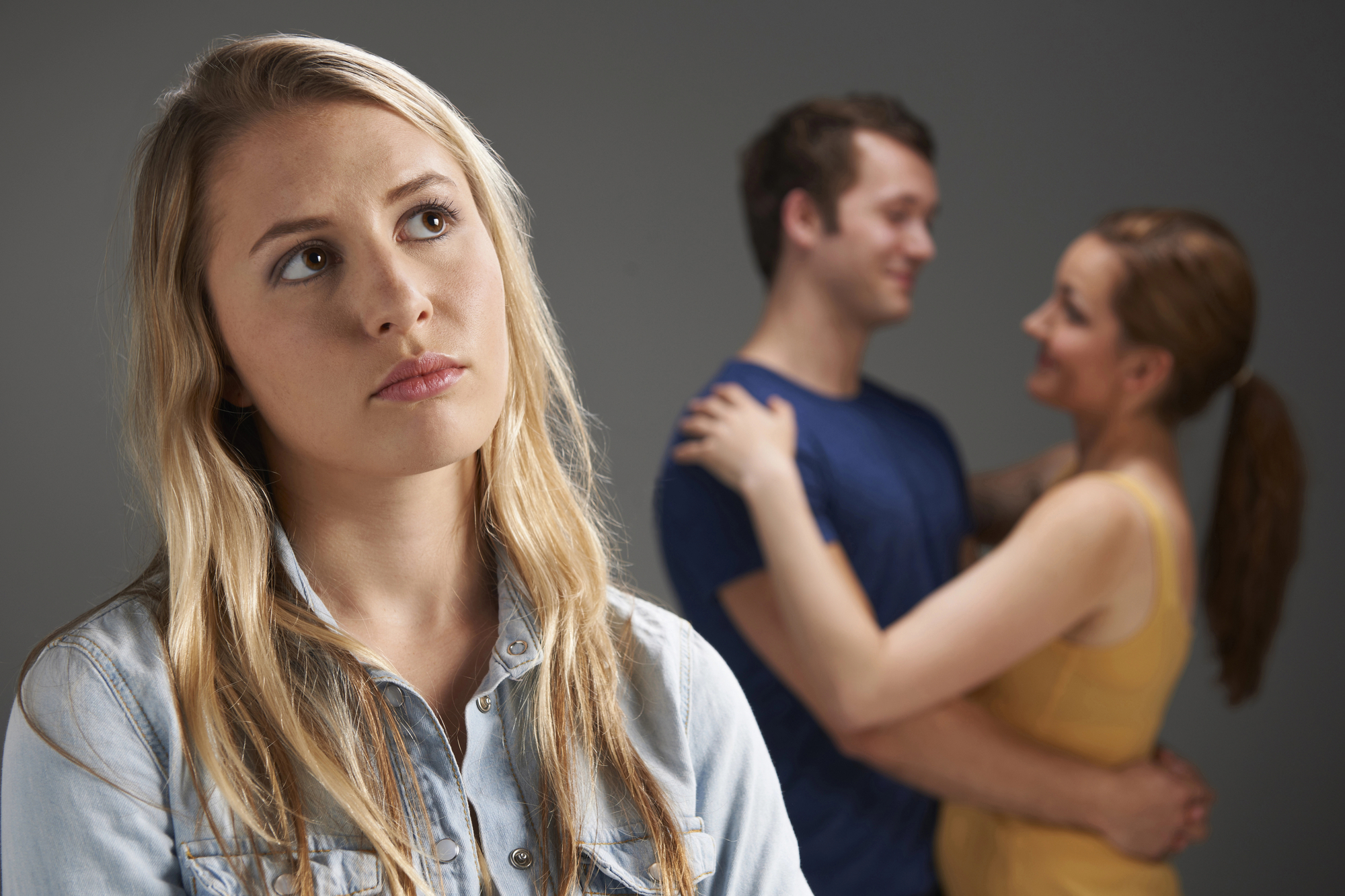 A woman with long blonde hair and a blue shirt looks upset in the foreground. In the background, a couple is embracing and smiling at each other. The woman's facial expression suggests feelings of sadness or jealousy.