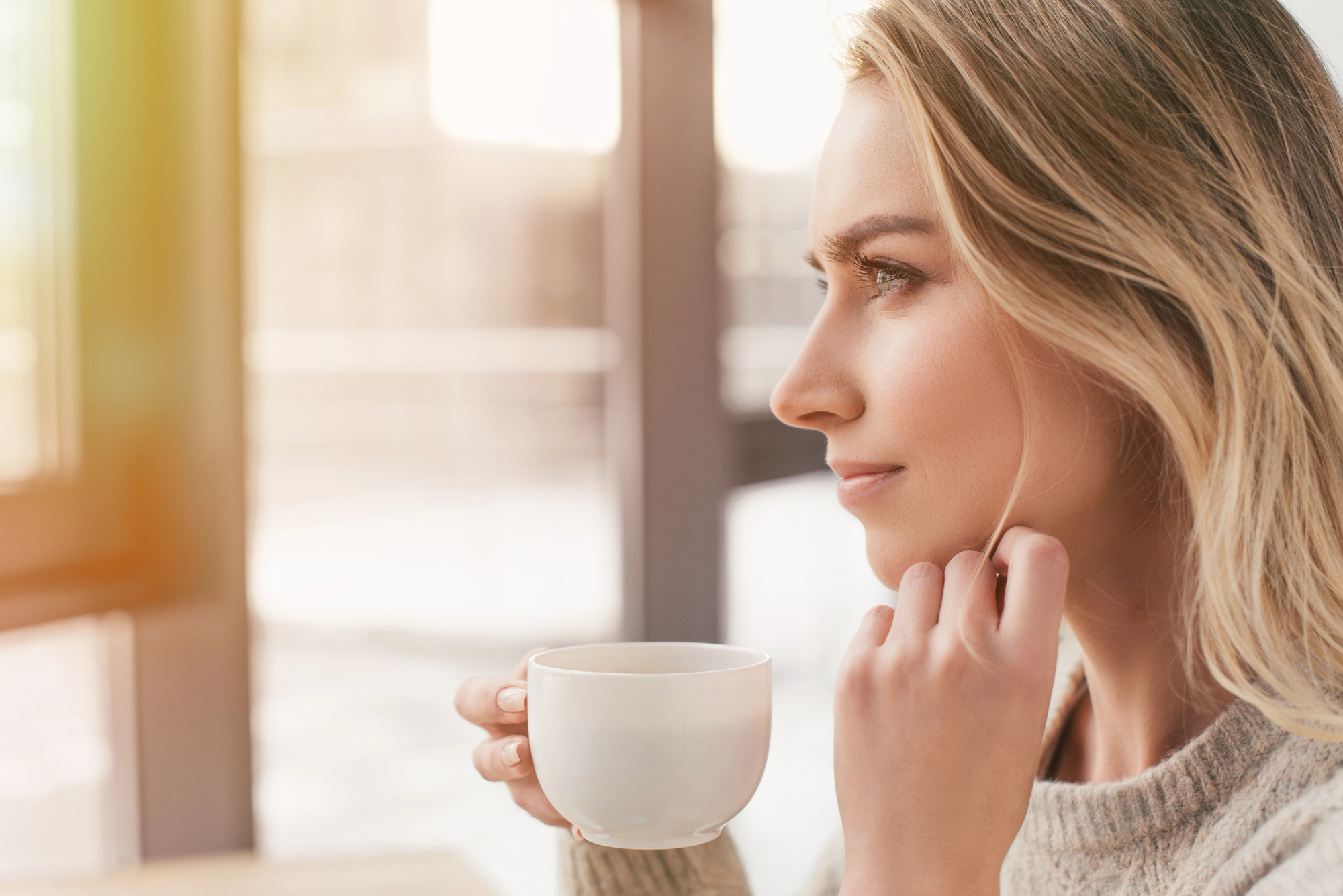 A blonde woman in a light sweater enjoys a warm drink from a white mug while gazing thoughtfully out of a window. Sunlight streams in, creating a serene and cozy atmosphere.