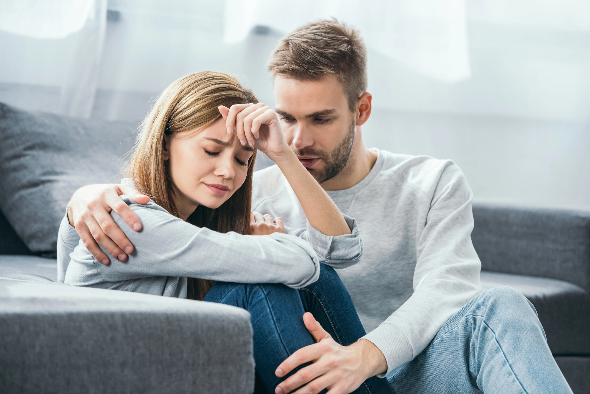 A man with a beard and short hair is sitting on a couch, comforting a woman with long brown hair who seems distressed. The woman has her eyes closed and is resting her forehead on her hand while the man has his arm around her shoulders. They both are wearing casual clothes.