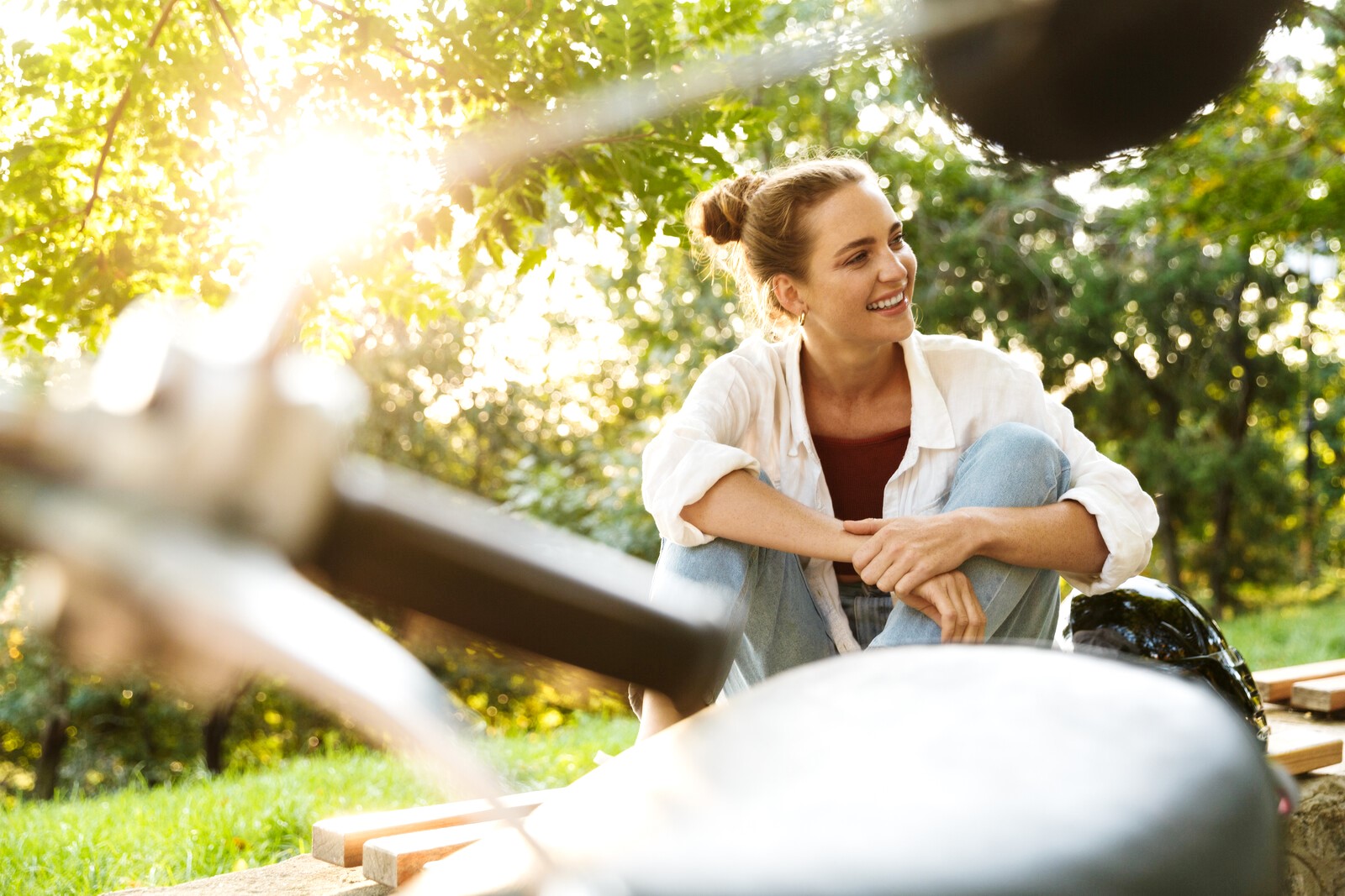 A woman with her hair in a bun sits outdoors under the trees, smiling and relaxed. She is wearing a white shirt over a dark top and light blue jeans. The sun is shining through the leaves, creating a warm, peaceful atmosphere. Nearby, there are blurred objects in the foreground.