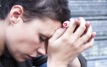 A person with short, dark hair and red nail polish is resting their forehead on their clasped hands, appearing deep in thought or distressed, against a blurred background of horizontal lines.