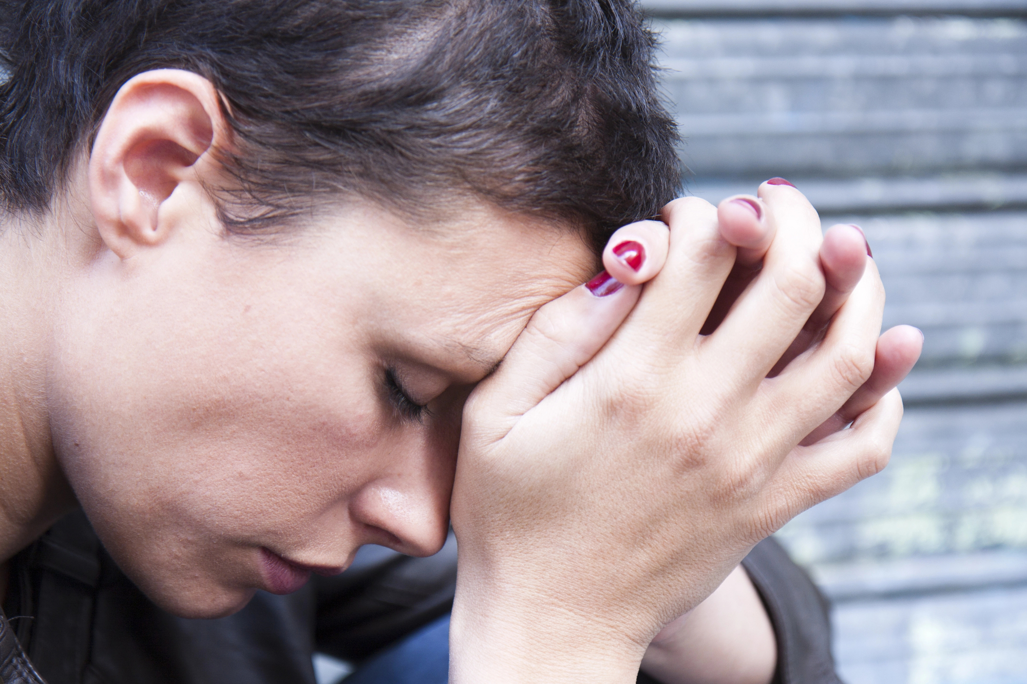 A person with short, dark hair and red nail polish is resting their forehead on their clasped hands, appearing deep in thought or distressed, against a blurred background of horizontal lines.