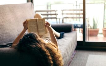 A person with curly hair lies on a gray couch, holding an open book above their face, reading. The room is well-lit by natural light coming through a sliding glass door, leading to a balcony with potted plants and outdoor furniture in the background.