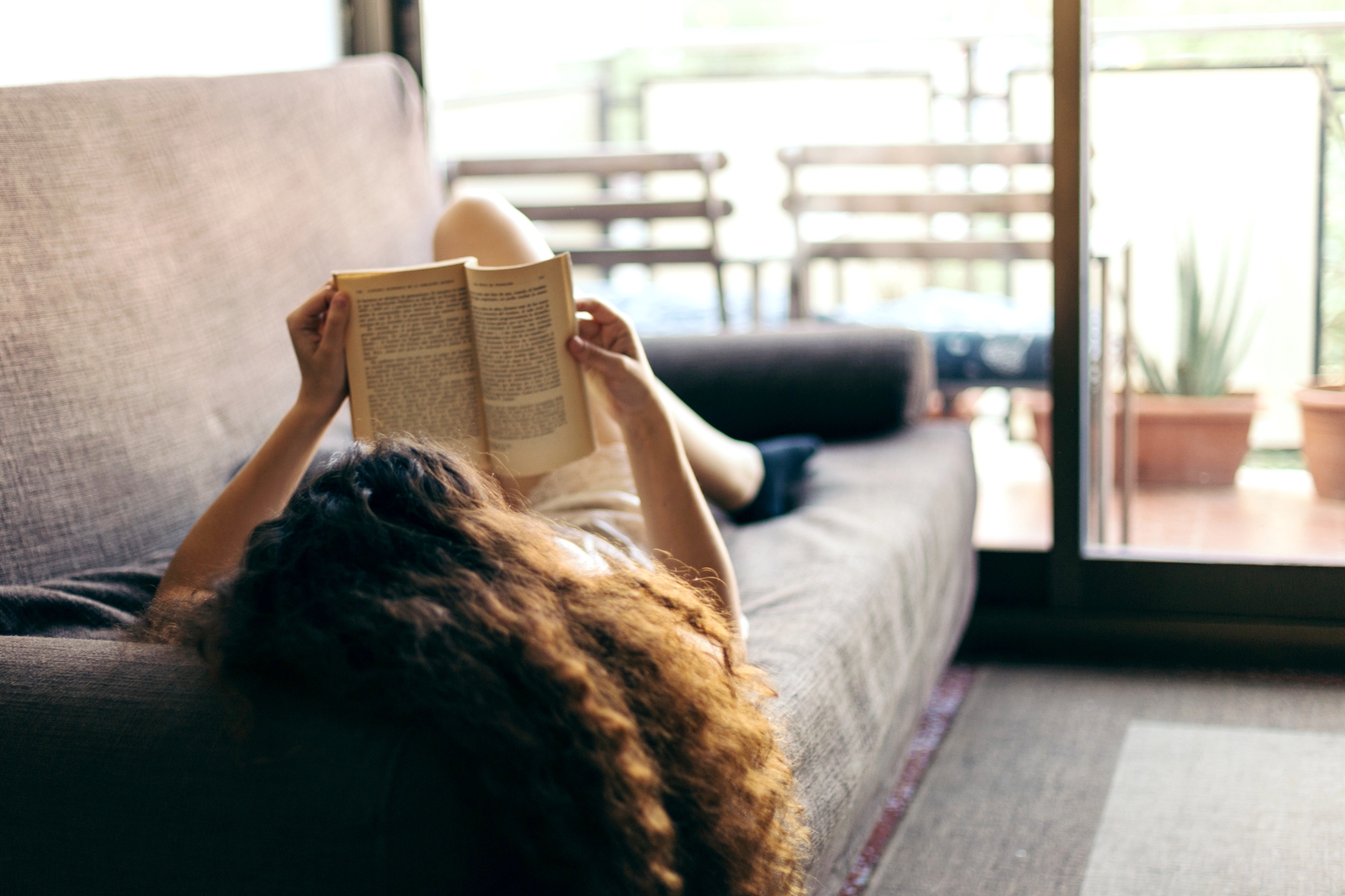 A person with curly hair lies on a gray couch, holding an open book above their face, reading. The room is well-lit by natural light coming through a sliding glass door, leading to a balcony with potted plants and outdoor furniture in the background.