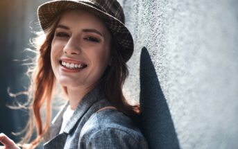 A person with long brown hair and wearing a hat smiles while leaning against a textured, gray wall. The individual is dressed in a gray blazer, and sunlight is softly illuminating the scene.