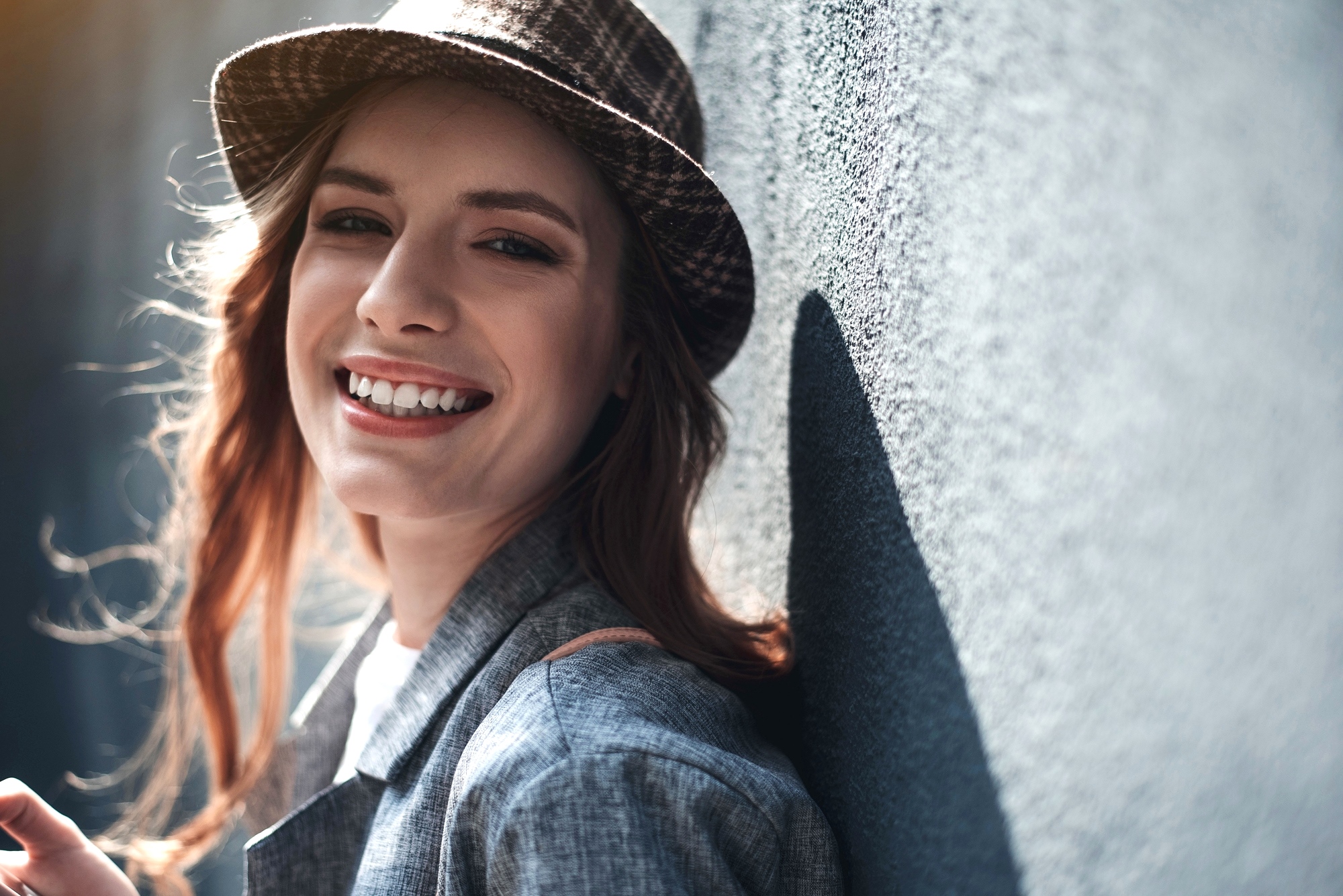 A person with long brown hair and wearing a hat smiles while leaning against a textured, gray wall. The individual is dressed in a gray blazer, and sunlight is softly illuminating the scene.