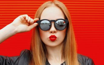 A person with long blonde hair is wearing black sunglasses and a black leather jacket. They are puckering their lips for a playful kiss against a bright red striped background. The reflection in the sunglasses shows an urban street scene.