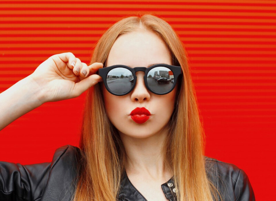 A person with long blonde hair is wearing black sunglasses and a black leather jacket. They are puckering their lips for a playful kiss against a bright red striped background. The reflection in the sunglasses shows an urban street scene.