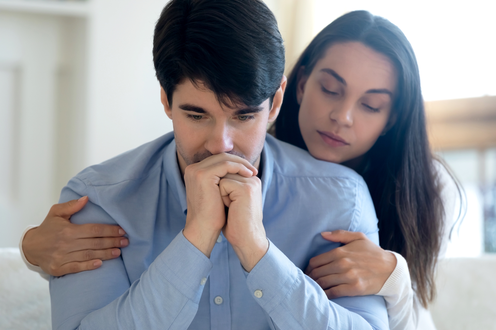 A person in a light blue shirt sits with a thoughtful expression and hands clasped near their face. Another person with long hair leans in from behind, gently embracing them and offering comfort. The scene suggests a moment of emotional support and empathy.