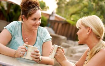 Two women are sitting outdoors at a table, engaged in conversation while holding mugs. Both are casually dressed and appear relaxed. Greenery is visible in the background, suggesting a garden or park setting. The atmosphere seems casual and friendly.