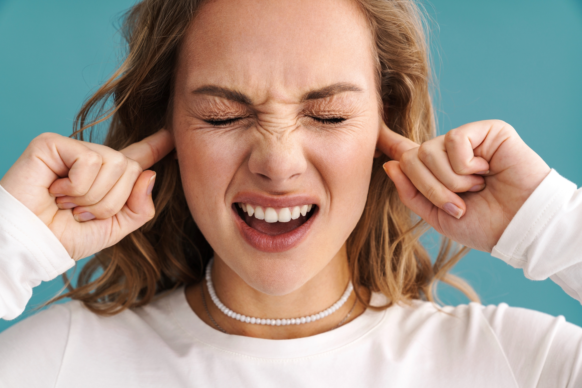 A woman with blonde hair, wearing a white shirt and a beaded necklace, has her eyes closed and is grimacing. She is covering her ears with both index fingers. The background is a solid teal color.