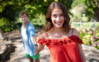 A woman in a red off-the-shoulder top smiles at the camera while being playfully pulled forward by a man in a blue shirt and green shorts in an outdoor setting with trees and greenery.