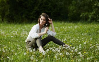 A woman sits cross-legged in a lush field of dandelions. She is smiling gently with her eyes closed, holding a dandelion close to her face. She wears a white shirt and dark pants. The background is filled with greenery, suggesting a serene and peaceful environment.