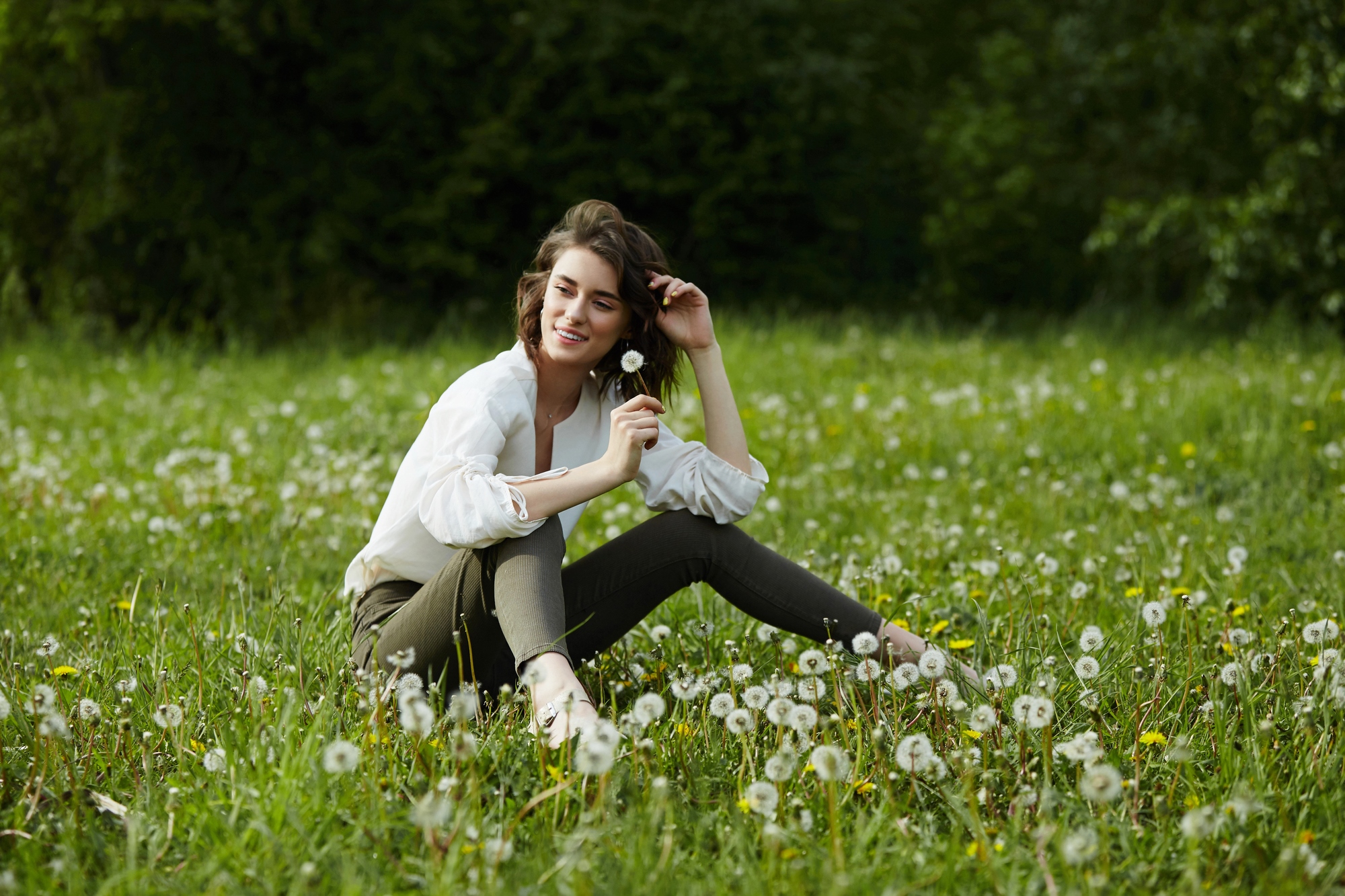 A woman sits cross-legged in a lush field of dandelions. She is smiling gently with her eyes closed, holding a dandelion close to her face. She wears a white shirt and dark pants. The background is filled with greenery, suggesting a serene and peaceful environment.