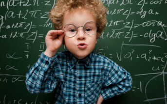 A young child with curly hair, wearing round glasses and a blue plaid shirt, stands in front of a chalkboard filled with mathematical equations. The child is confidently posing with one hand on hip and the other adjusting the glasses, looking directly at the camera.