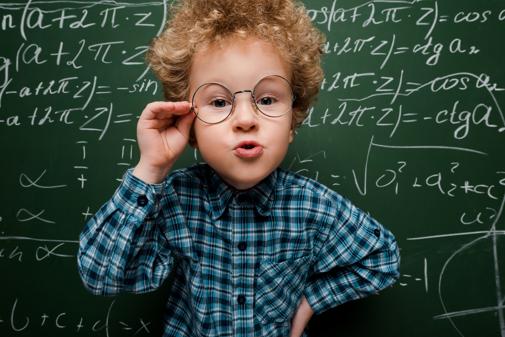 A young child with curly hair, wearing round glasses and a blue plaid shirt, stands in front of a chalkboard filled with mathematical equations. The child is confidently posing with one hand on hip and the other adjusting the glasses, looking directly at the camera.