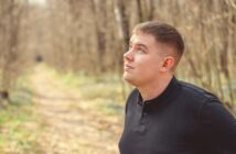 A young man with short hair, wearing a black long-sleeve shirt, gazes upward while standing on a sunlit forest path. Trees with bare branches line the trail, receding into the background. The setting appears serene and contemplative.