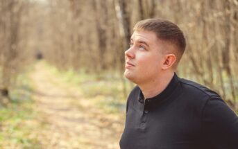 A young man with short hair, wearing a black long-sleeve shirt, gazes upward while standing on a sunlit forest path. Trees with bare branches line the trail, receding into the background. The setting appears serene and contemplative.