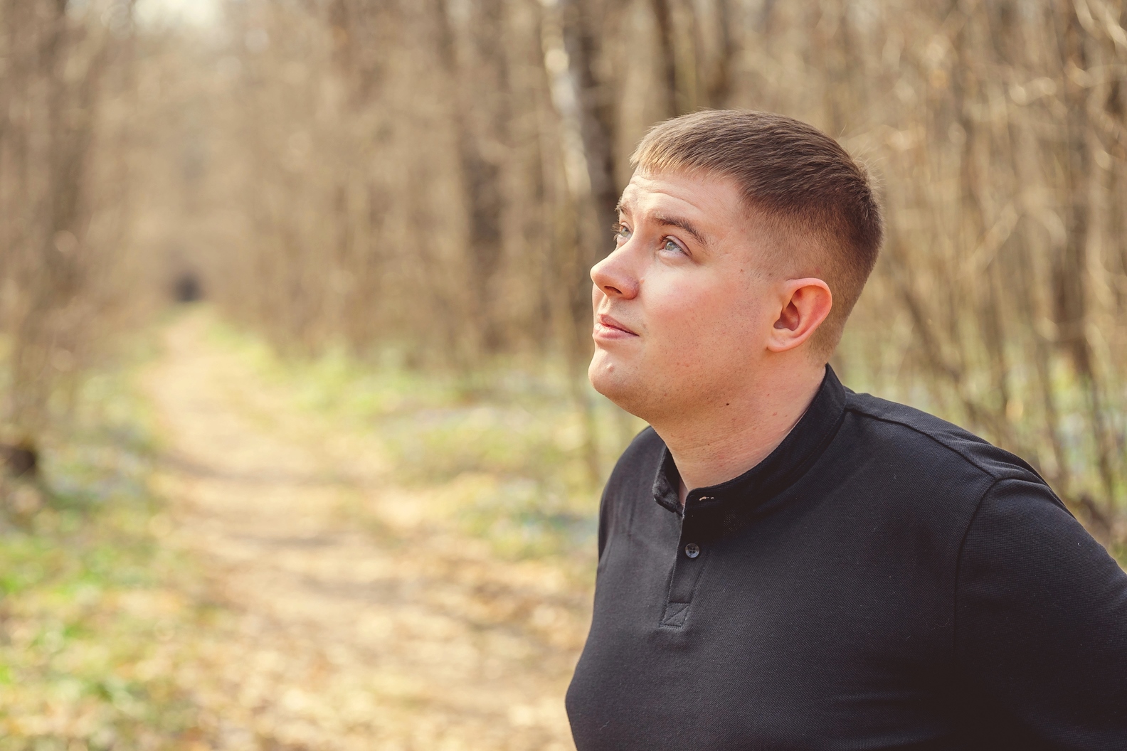 A young man with short hair, wearing a black long-sleeve shirt, gazes upward while standing on a sunlit forest path. Trees with bare branches line the trail, receding into the background. The setting appears serene and contemplative.