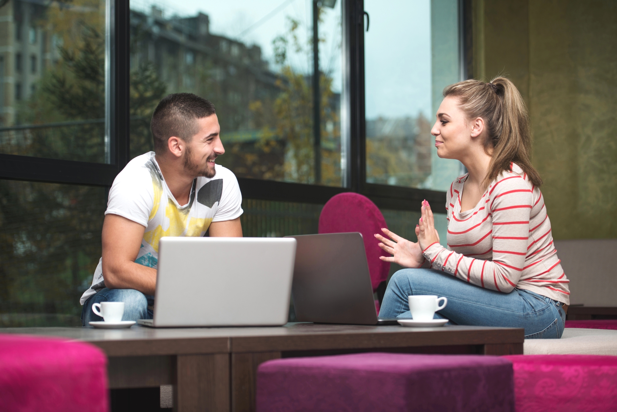 Two people sit across from each other at a table with laptops and coffee cups. They are engaged in conversation and smiling. The background includes large windows revealing an urban, green scenery outside. Both seem relaxed and casual in their attire.